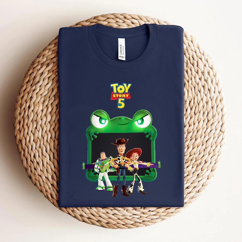 May include: Navy blue t-shirt featuring a green frog-shaped frame with the text "TOY STORY 5". Inside the frame are cartoon characters Woody, Buzz Lightyear, and Jessie. The shirt is displayed on a woven, circular surface.
