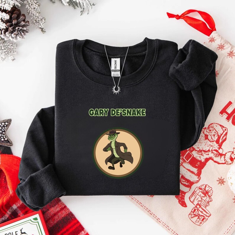 May include: Black sweatshirt with the text "GARY DE'SNAKE" and a cartoon snake in a hat and coat. The snake is inside a circular frame. The sweatshirt is folded on a white surface, with festive decorations visible in the background.