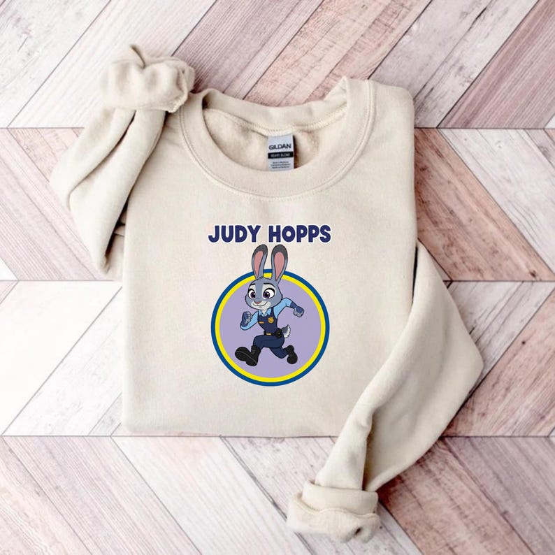 May include: A cream-colored sweatshirt featuring the text "JUDY HOPPS" above a cartoon image of Judy Hopps, a rabbit character in a police uniform, inside a blue and yellow circle. The sweatshirt is folded on a wooden surface.