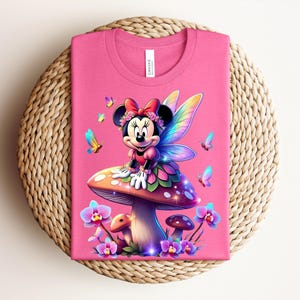 May include: A pink t-shirt featuring a cartoon Minnie Mouse dressed as a fairy, sitting on a mushroom. The design includes colorful butterflies and flowers. The shirt is folded on a woven, circular surface.
