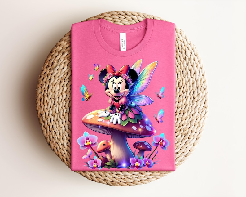 May include: A pink t-shirt featuring a cartoon Minnie Mouse dressed as a fairy, sitting on a mushroom. The design includes colorful butterflies and flowers. The shirt is folded on a woven, circular surface.