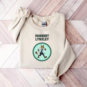 May include: Cream-colored sweatshirt with the text "PAWBERT LYNXLEY" above a circular graphic. The graphic features a cartoon lynx wearing a lab coat and walking. The sweatshirt has long sleeves and a crew neck.
