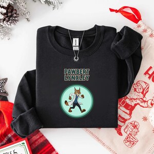 May include: Black crewneck sweatshirt featuring the text "PAWBERT LYNXLEY" above a cartoon lynx character in a lab coat. The lynx is inside a light green circle. A silver necklace is visible.