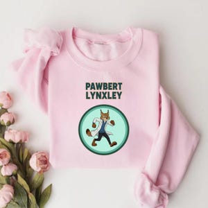 May include: A light pink sweatshirt with the text "PAWBERT LYNXLEY" above a circular graphic. The graphic features a cartoon lynx wearing a lab coat and pants, set against a teal green background.