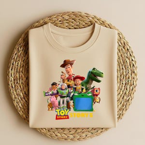 K&ouml;nnte beinhalten: Ein beiges T-Shirt mit einer farbenfrohen Grafik von Charakteren aus dem Animationsfilm "Toy Story". Das Design umfasst Woody, Buzz Lightyear, Jessie, Rex und andere Charaktere, mit dem Logo "Toy Story 5".