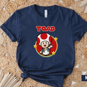 Może przedstawiać: Granatowa koszulka z kresk&oacute;wkową ilustracją Toada z Super Mario Bros. Postać znajduje się w czerwonym okręgu z ż&oacute;łtymi gwiazdami, a nadruk "TOAD" jest w czerwonych literach.
