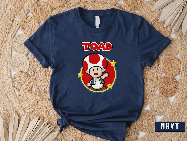 Może przedstawiać: Granatowa koszulka z kresk&oacute;wkową ilustracją Toada z Super Mario Bros. Postać znajduje się w czerwonym okręgu z ż&oacute;łtymi gwiazdami, a nadruk "TOAD" jest w czerwonych literach.