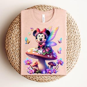 May include: A light pink t-shirt featuring a colorful illustration of Minnie Mouse as a fairy, sitting on a mushroom. The design includes butterflies and flowers. The shirt is folded on a woven, circular surface.