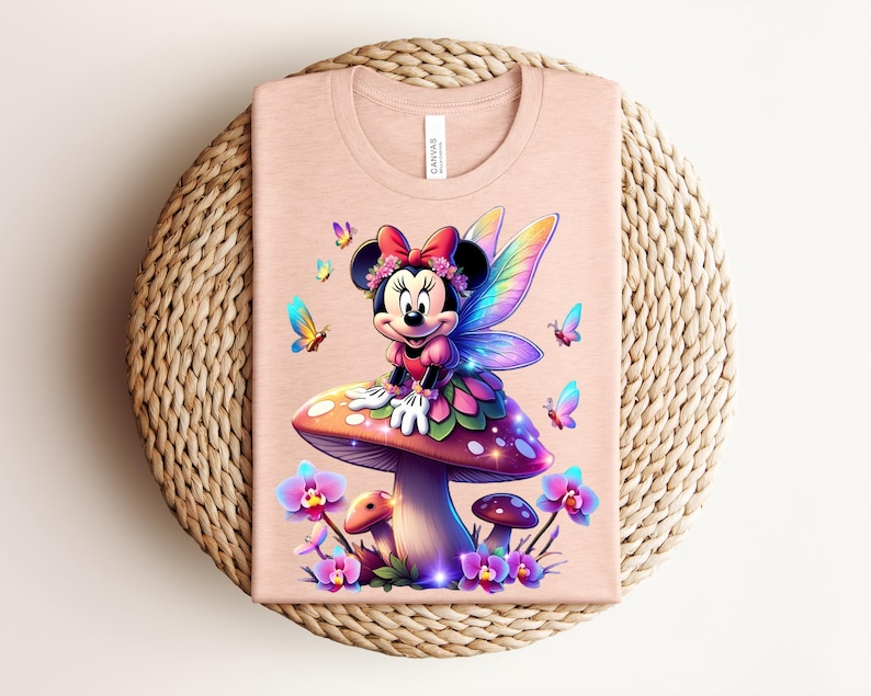 May include: A light pink t-shirt featuring a colorful illustration of Minnie Mouse as a fairy, sitting on a mushroom. The design includes butterflies and flowers. The shirt is folded on a woven, circular surface.