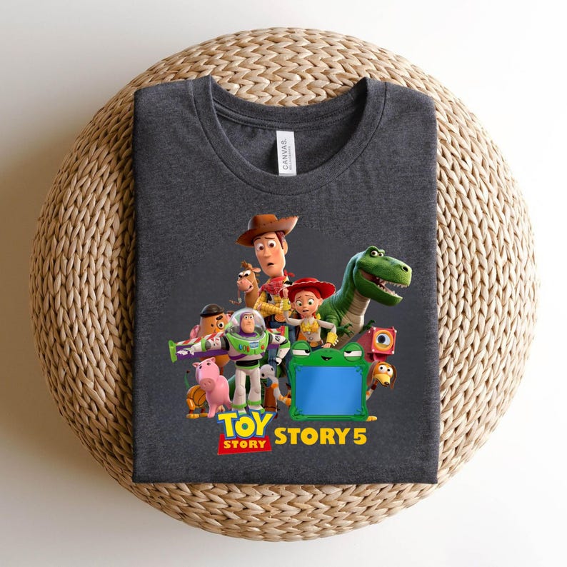 K&ouml;nnte beinhalten: Dunkelgraues T-Shirt mit einer farbenfrohen Grafik von Charakteren aus dem Animationsfilm "Toy Story". Das Design zeigt Woody, Buzz Lightyear, Rex und andere Charaktere mit dem Text "Toy Story 5" in Gelb und Rot.