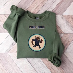 May include: An olive green sweatshirt with the text "CHIEF BOGO" above a circular graphic. The graphic features a cartoon bull in a police uniform running inside a blue and tan circle.