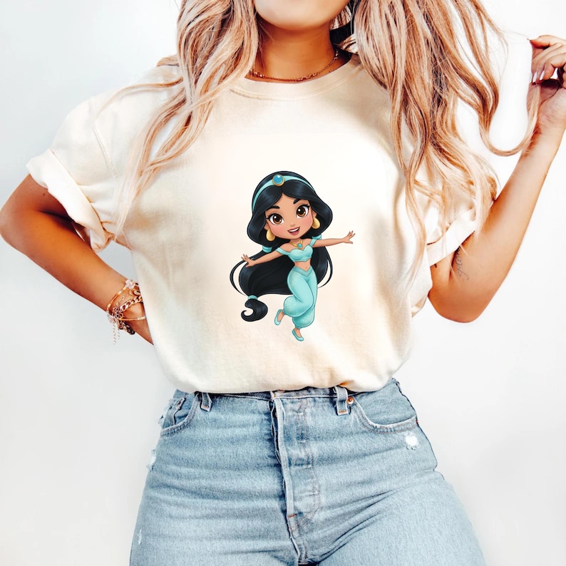 Puede incluir: Camiseta color crema con una ilustraci&oacute;n de dibujos animados de una princesa con un atuendo turquesa, cabello negro y aretes dorados. La princesa est&aacute; en una pose alegre con los brazos extendidos. La camiseta se combina con jeans azules.