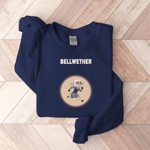 Op de afbeelding: Marineblauwe sweatshirt met het woord "BELLWETHER" in witte blokletters. Een cartoon schaap met bril en blauwe jurk staat in een cirkelvormig ontwerp. De sweatshirt is gemaakt van een zacht materiaal.