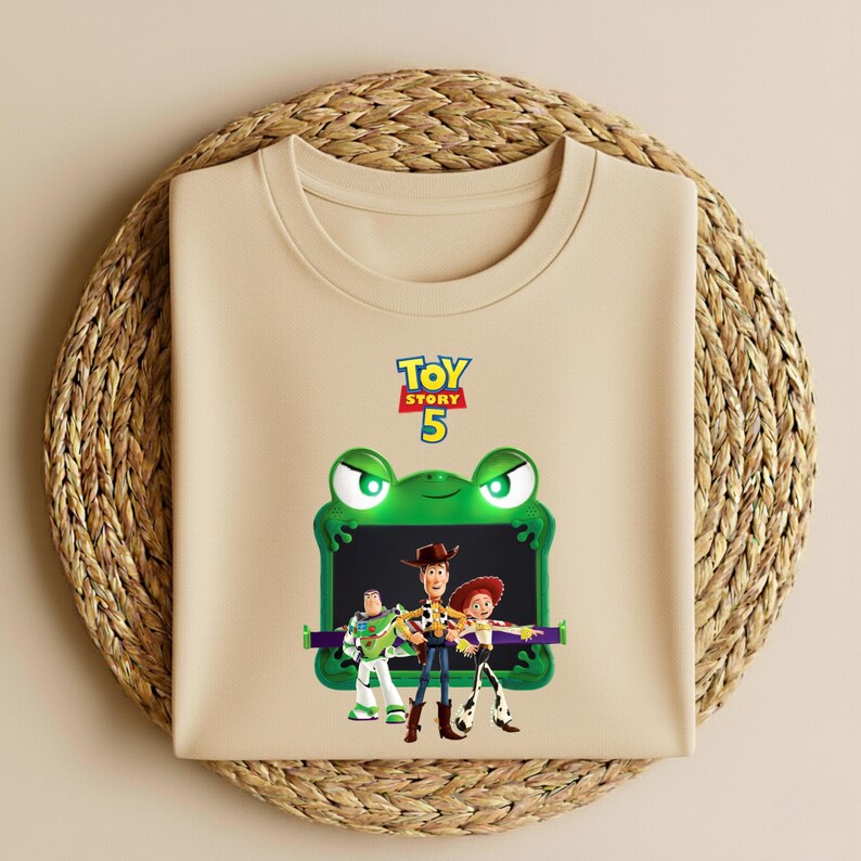May include: A beige t-shirt featuring a graphic of characters from the movie Toy Story 5. The graphic includes Buzz Lightyear, Woody, and Jessie, framed by a green frog-like design. The words "Toy Story 5" are at the top.