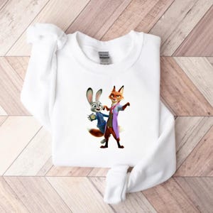 May include: White sweatshirt featuring a colorful graphic of two cartoon characters, a rabbit and a fox, in a friendly pose. The rabbit wears a blue jacket, and the fox wears a purple coat. The sweatshirt is made of a soft material.