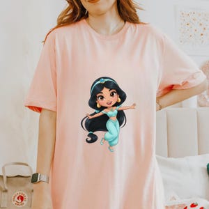 Puede incluir: Camiseta rosa claro con una ilustraci&oacute;n de dibujos animados de la princesa Jasmine. Lleva un atuendo turquesa, pelo negro y pendientes dorados. La princesa est&aacute; en una pose din&aacute;mica con los brazos extendidos.