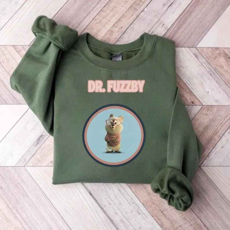 May include: An olive green sweatshirt with the text "DR. FUZZBY" in pink. The design includes a cartoon character in a circle. The sweatshirt is folded on a wooden surface with a chevron pattern.
