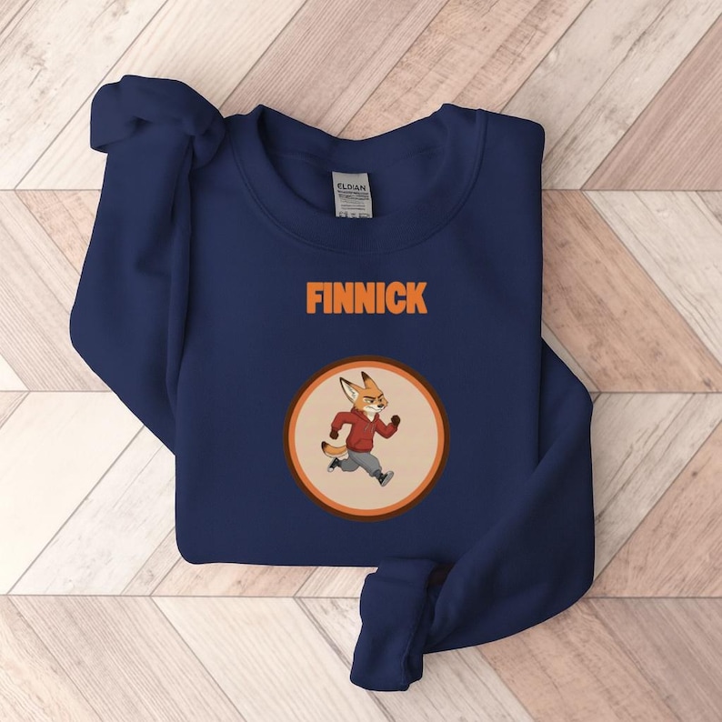 Op de afbeelding: Marineblauwe sweatshirt met de naam "FINNICK" in oranje blokletters. Een cirkelvormige afbeelding toont een cartoonvos in een rode jas en grijze broek. De vos rent. De sweatshirt is gemaakt van een zacht materiaal.