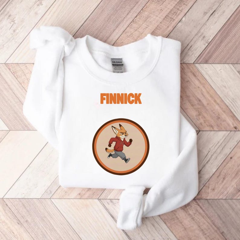 Op de afbeelding: Witte sweatshirt met de naam "FINNICK" in oranje blokletters boven een cirkelvormige afbeelding. De afbeelding toont een cartoonvos in een rode jas en grijze broek, rennend in een bruine en oranje cirkel.