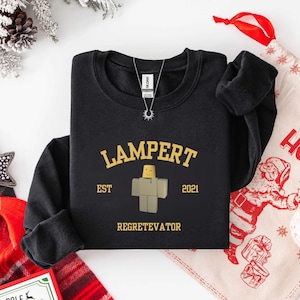 May include: Black sweatshirt with the word "LAMPERT" in gold lettering above a block figure and the text "EST 2021 REGETEVATOR". A silver necklace with a sun pendant hangs from the collar.