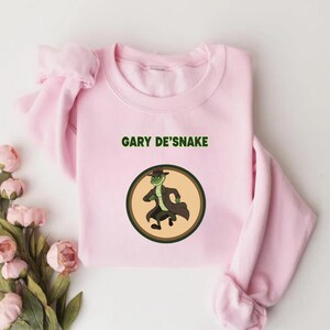 May include: A pale pink sweatshirt with the words "GARY DE'SNAKE" in green. Below the text is a circular graphic of a cartoon snake wearing a hat and trench coat. The snake is green, set on a tan background.
