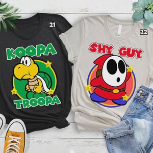 Może przedstawiać: Dwie koszulki z grafikami postaci z kresk&oacute;wek. Jedna czarna koszulka przedstawia wz&oacute;r Koopa Troopa w kolorze zielonym i ż&oacute;łtym. Druga to beżowa koszulka z grafiką Shy Guy w kolorze czerwonym, białym i niebieskim. Obie koszulki mają nadrukowane imię postaci.
