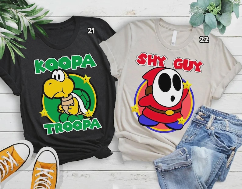 Może przedstawiać: Dwie koszulki z grafikami postaci z kresk&oacute;wek. Jedna czarna koszulka przedstawia wz&oacute;r Koopa Troopa w kolorze zielonym i ż&oacute;łtym. Druga to beżowa koszulka z grafiką Shy Guy w kolorze czerwonym, białym i niebieskim. Obie koszulki mają nadrukowane imię postaci.
