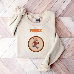 Op de afbeelding: Cr&egrave;mekleurige sweatshirt met de naam "FINNICK" in oranje blokletters. Een cirkelvormige afbeelding toont een cartoonvos in een rode jas en grijze broek. De vos rent in een bruine en oranje cirkel.
