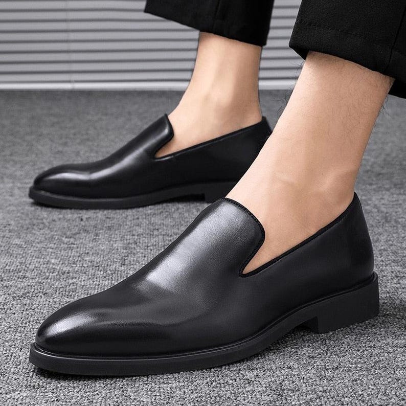 May include: Black leather slip-on shoes with a low heel. The shoes have a sleek, polished appearance and a classic design. The shoes are shown on a gray carpet.