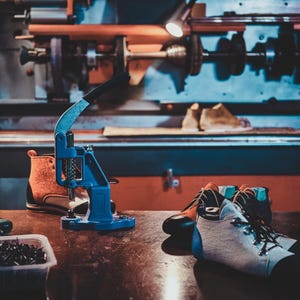 Pode incluir: Oficina de sapateiro com diversas ferramentas e materiais. Uma prensa azul, botas inacabadas e um par de botas de caminhada brancas e pretas numa mesa de madeira. Uma m&aacute;quina grande ao fundo.
