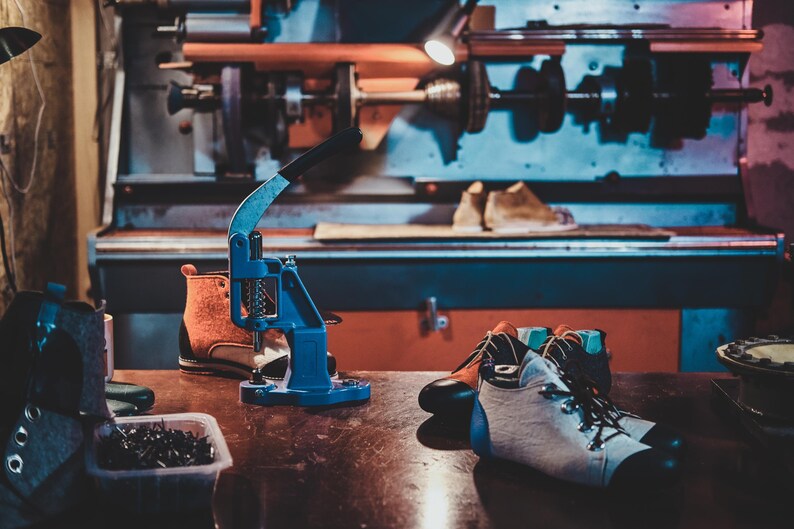 Pode incluir: Oficina de sapateiro com diversas ferramentas e materiais. Uma prensa azul, botas inacabadas e um par de botas de caminhada brancas e pretas numa mesa de madeira. Uma m&aacute;quina grande ao fundo.