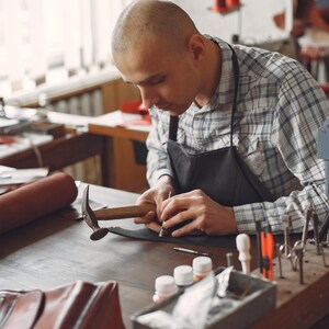 Pode incluir: Uma pessoa trabalha em artigos de couro numa oficina. Ele est&aacute; usando um martelo e outras ferramentas em uma pe&ccedil;a de couro marrom escuro. Um rolo de couro marrom e uma bolsa de couro est&atilde;o sobre a mesa. A pessoa est&aacute; usando uma camisa xadrez e um avental preto.