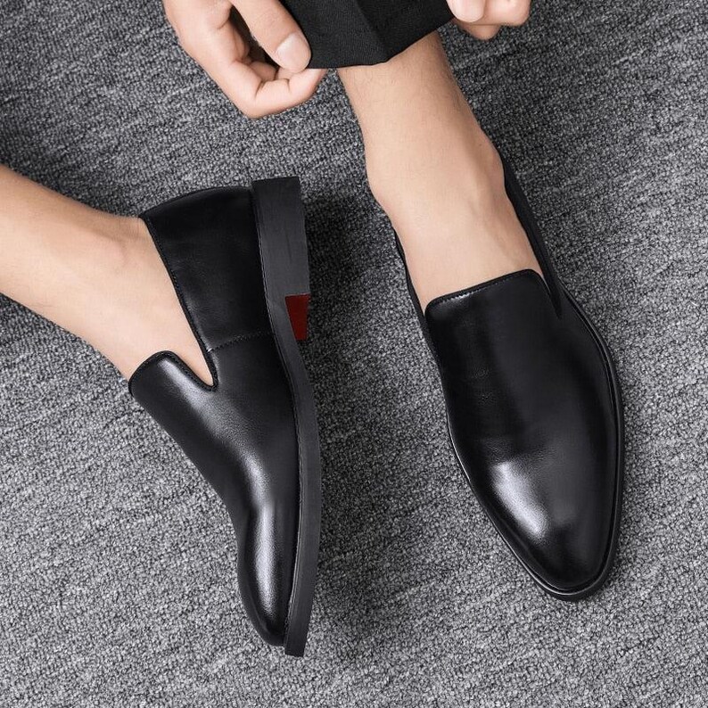 May include: Close-up of two black leather slip-on shoes. The shoes have a sleek, polished appearance with a pointed toe design. The soles are black with a red accent. The shoes are on a gray surface.