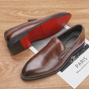 May include: A pair of brown leather slip-on shoes with a glossy finish. The soles are black with a red accent. One shoe is positioned upside down, revealing the red sole. The other shoe is right-side up, with a pointed toe. A paper with the word "PARIS" is visible.