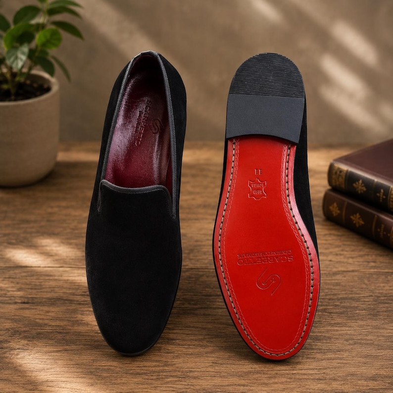 May include: Black velvet loafers with a red leather sole. The shoes have a classic design with a rounded toe and a slip-on style. The sole is stitched with a contrasting thread. The interior is a deep burgundy color.