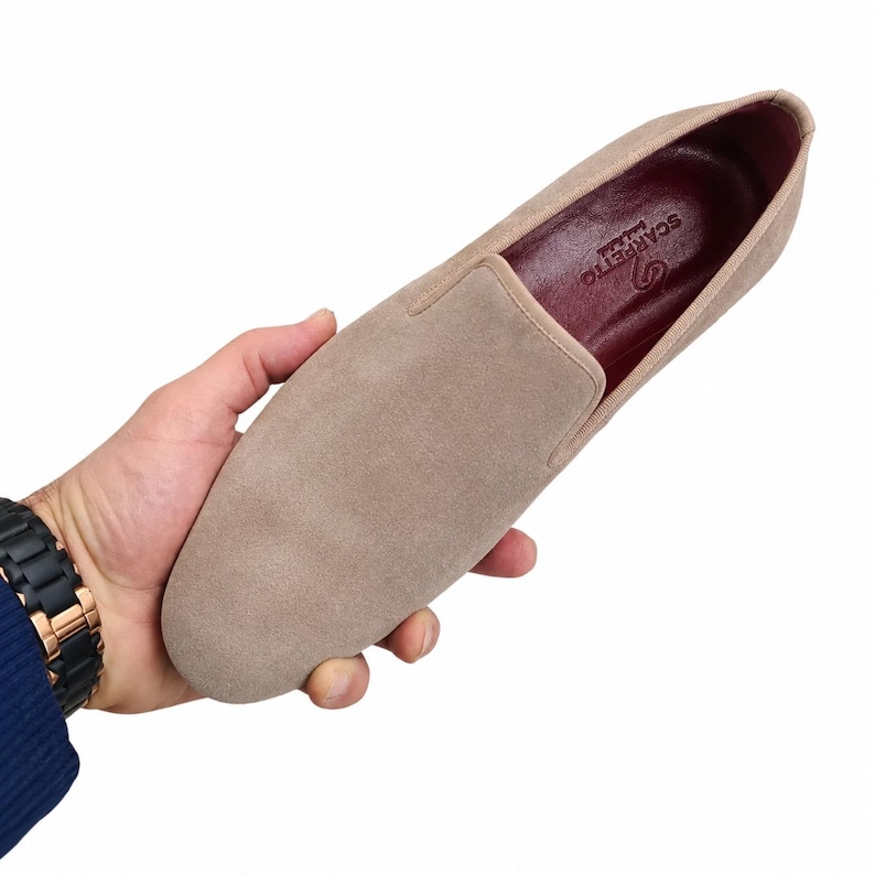 May include: A beige suede slip-on shoe with a rounded toe. The shoe has a dark red interior and a light beige trim. The brand name "Scarpetto" is visible inside the shoe. The shoe is held in a hand.