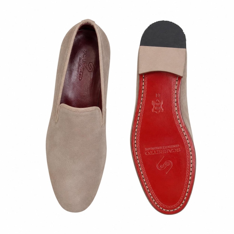 May include: Two beige suede slip-on shoes. One shoe is shown from the top, the other from the sole. The sole is red with white stitching around the edge. The brand name is visible on the sole.