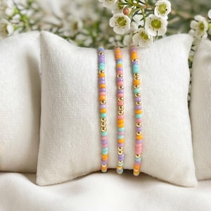 May include: Three sets of beaded bracelets in pastel colors, including orange, yellow, blue, and purple, are displayed on white pillows. Each set includes a gold chain bracelet. White flowers are in the background, creating a soft, feminine aesthetic.