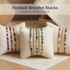 May include: Three sets of beaded bracelets displayed on cream-colored velvet pillows. The bracelets feature various color combinations of small glass beads and metallic gold-tone beads. Text at the top reads: Football Bracelet Stacks Custom Team Colors.