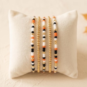 May include: A set of five beaded bracelets arranged vertically. Three bracelets feature a repeating pattern of small orange, black, white, and metallic gold beads. Two bracelets consist entirely of small, uniform metallic gold beads.
