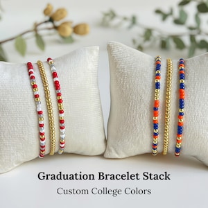 May include: Two sets of beaded bracelets in custom college colors, displayed on white pillows. One set features red, white, and gold beads, while the other showcases blue, orange, and gold beads. The text reads "Graduation Bracelet Stack" and "Custom College Colors."