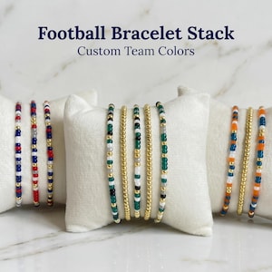 May include: A stack of beaded bracelets in various team colors, displayed on white pillows. The bracelets feature gold beads and colorful accents, with text that reads "Football Bracelet Stack Custom Team Colors."