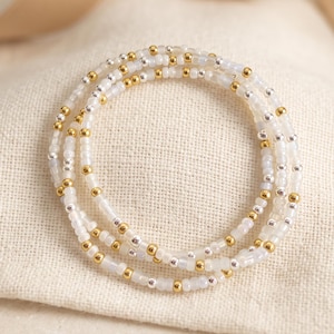 May include: A multi-strand beaded bracelet featuring a repeating pattern of translucent white glass beads interspersed with small, polished gold-tone and silver-tone metallic spherical beads arranged in a circular formation.