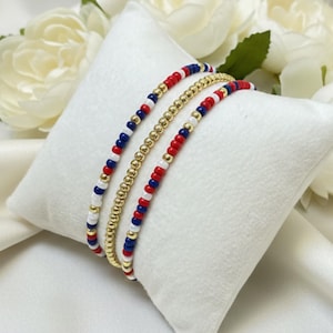 May include: Three beaded bracelets in red, white, blue, and gold colors are displayed on a white pillow. The bracelets are made of small, round beads. The background features white flowers and a white satin fabric.