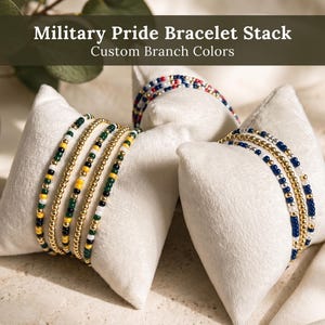May include: A collection of beaded bracelets displayed on two white velvet pillows. The bracelets feature small round beads in gold, white, green, yellow, blue, and black. The text Military Pride Bracelet Stack Custom Branch Colors is visible.