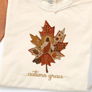 May include: An ivory t-shirt featuring a large faux patchwork maple leaf graphic. The leaf contains images of a figure, crosses, sunflowers, pumpkins, and the text Faith Hope Love. Below the leaf, the text autumn grace is printed in brown.