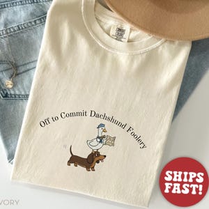 May include: Ivory-colored t-shirt with the phrase "Off to Commit Dachshund Foolery" in a curved font. The graphic features a dachshund dog with a goose wearing a sailor hat and holding a map. The shirt has a Comfort Colors tag.