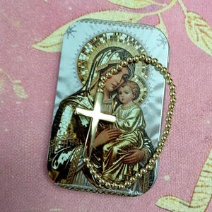 May include: A rectangular metal tin featuring a printed religious icon of a figure holding a child, overlaid with a gold-toned beaded rosary chain and a smooth, polished gold-toned cross pendant resting on the surface of the container.