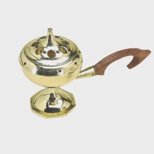 May include: A polished brass incense burner featuring a rounded, perforated lid with a central finial, a bowl-shaped body, and a hexagonal pedestal base. A curved, dark brown wooden handle is attached to the side of the metallic vessel.
