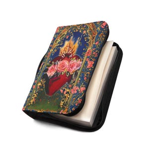 May include: A black zippered case with a decorative cover featuring a red heart with pink roses, flames, and a cross. The cover has a blue background with floral and gold accents. The case is partially open, revealing white pages.